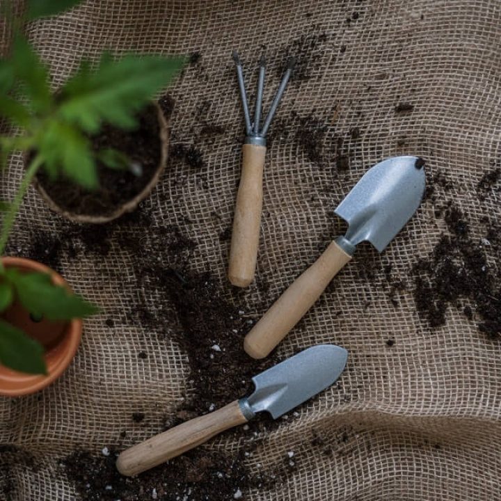 "Conjunto de herramientas de jardinería eco-friendly sobre un paño de arpillera: dos palas pequeñas y un rastrillo de mano con mangos de madera, acompañados de tierra esparcida, dos macetas emergentes y una bolsa de papel con tierra, evocando prácticas sostenibles y la conexión con la naturaleza en un hogar ecológico
